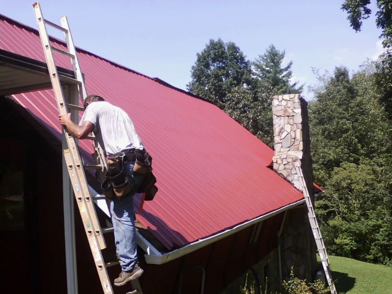Roofer on a ladder beside a red metal roof and stone chimney — Soffit & Fascia Repair by Roofing Brothers in Whipple
